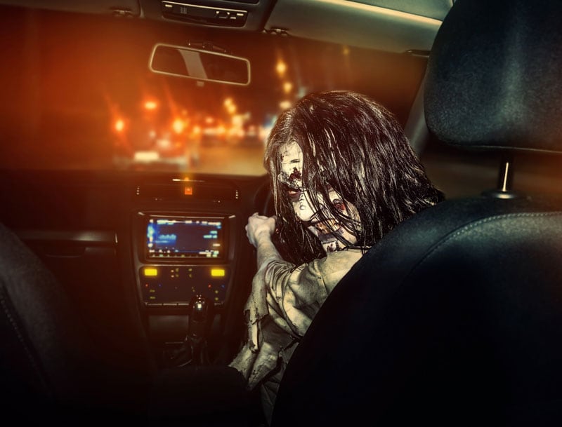 Zombie rides in a car at nighttime with a sinister grin